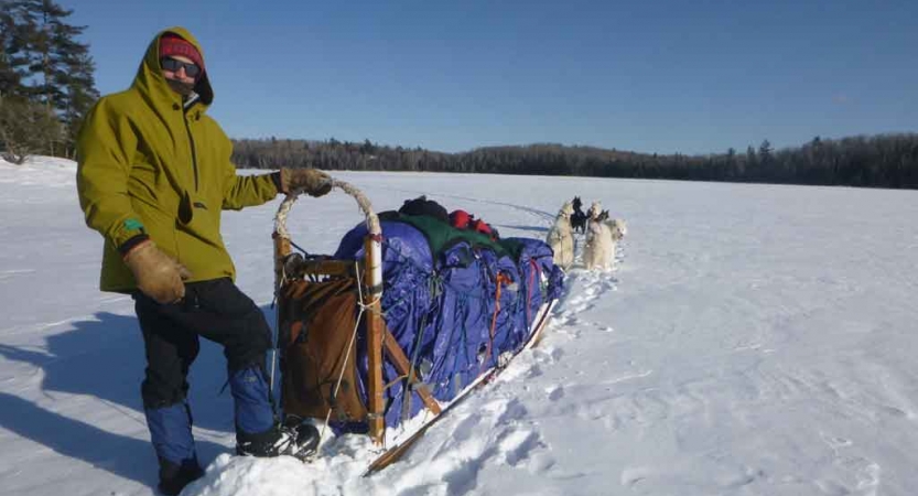 a gap year student poses with a dog sled in on a frozen, snowy lake lined with trees